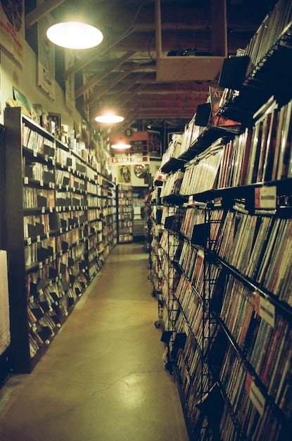 Interior of a bustling, dimly lit record store with shelves full of vinyl. Several young people are sifting through records, chatting, and enjoying the atmosphere.